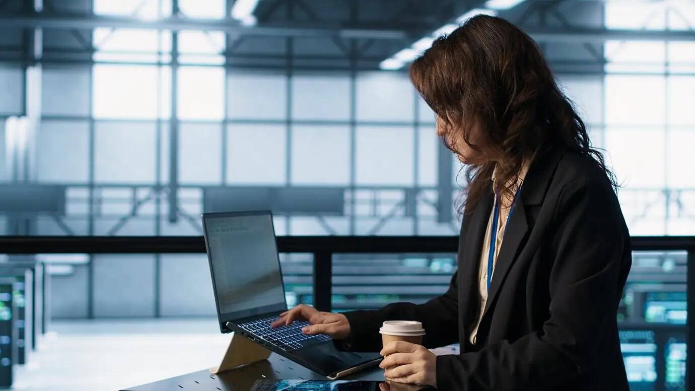 Mujer en un centro de datos usando un portátil para actualizar matrices de almacenamiento mientras sostiene una taza de café.